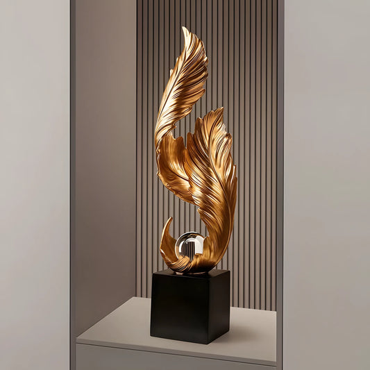 A modern decorative sculpture featuring two sweeping metallic gold leaf-like forms rising from a glossy black rectangular base, displayed on a shelf against a slatted wood and gray wall background.