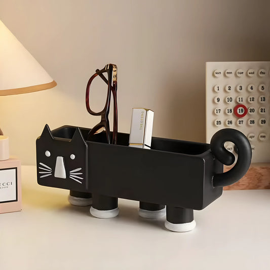 A black cat-shaped desk organizer with white facial features and white suction cup feet, holding eyeglasses and a lipstick tube, on a tabletop with a lamp and calendar in the background.