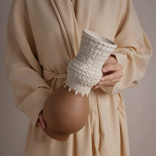 A person in a beige robe holds a sculptural ceramic vase with a textured cream upper section and a smooth tan lower section against a neutral beige background.