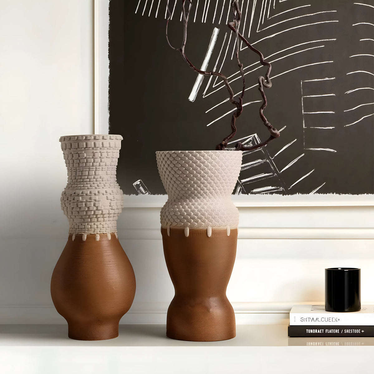 Two sculptural ceramic vases displayed on a white shelf in an interior setting, with a framed abstract artwork and a small black candle in the background.