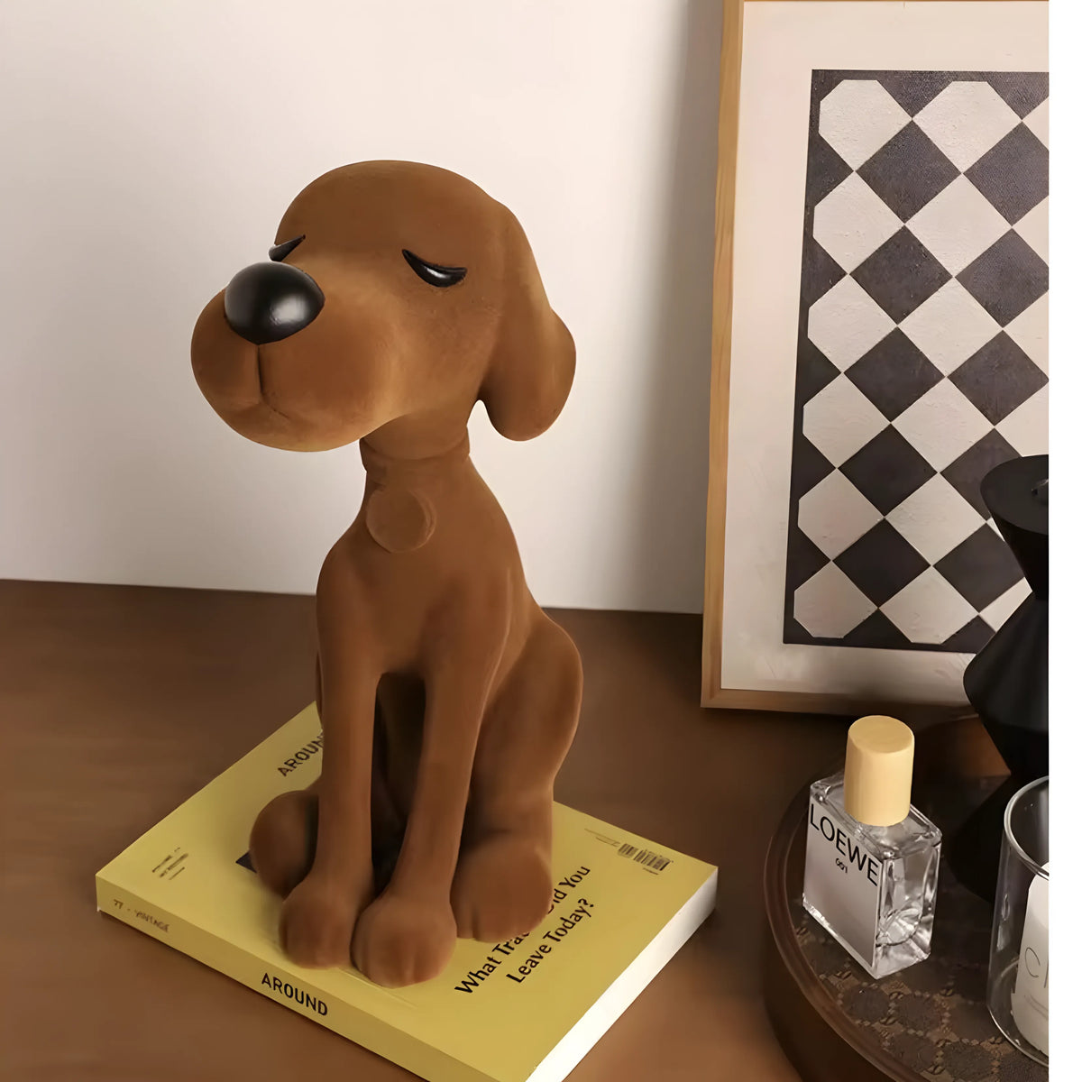 A brown plush dog figurine sitting on a yellow book on a wooden table, with a Loewe perfume bottle and a framed black-and-white geometric print in the background.