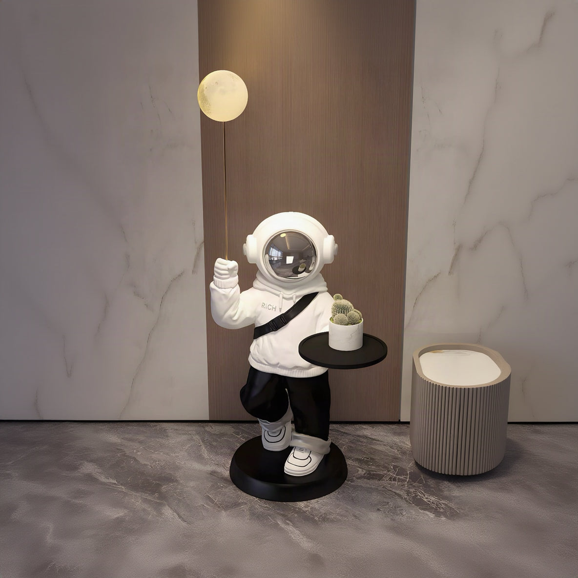 A whimsical astronaut figurine holding a glowing moon lamp on a string and a small potted cactus on a black side table, set in a modern interior with marble walls and flooring.