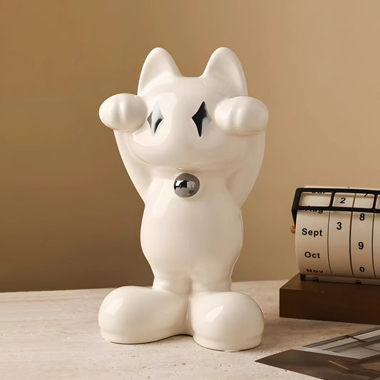 A glossy white ceramic cat figurine with a raised paw and a small silver bell at its neck, displayed on a tabletop next to a vintage-style calendar against a warm beige background.