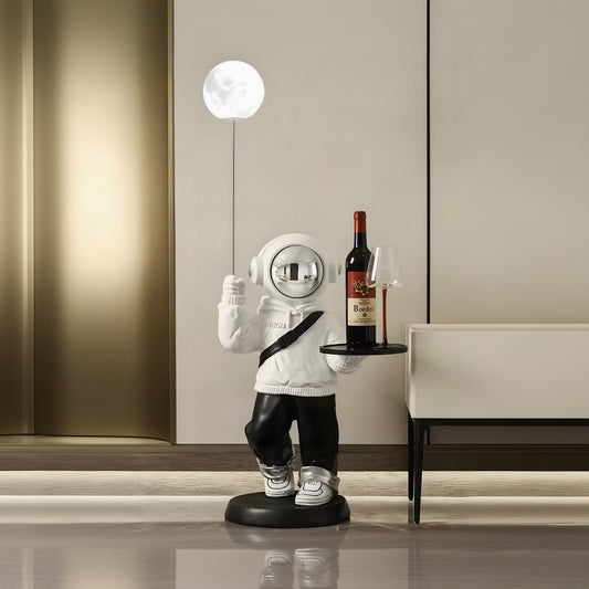 A whimsical astronaut-shaped floor lamp or decorative figure stands on a round black base in a modern interior. The figure is dressed in a white hoodie and black pants, holding a small tray with a wine bottle and a glass, while a glowing moon-shaped bulb hangs above on a thin cord.