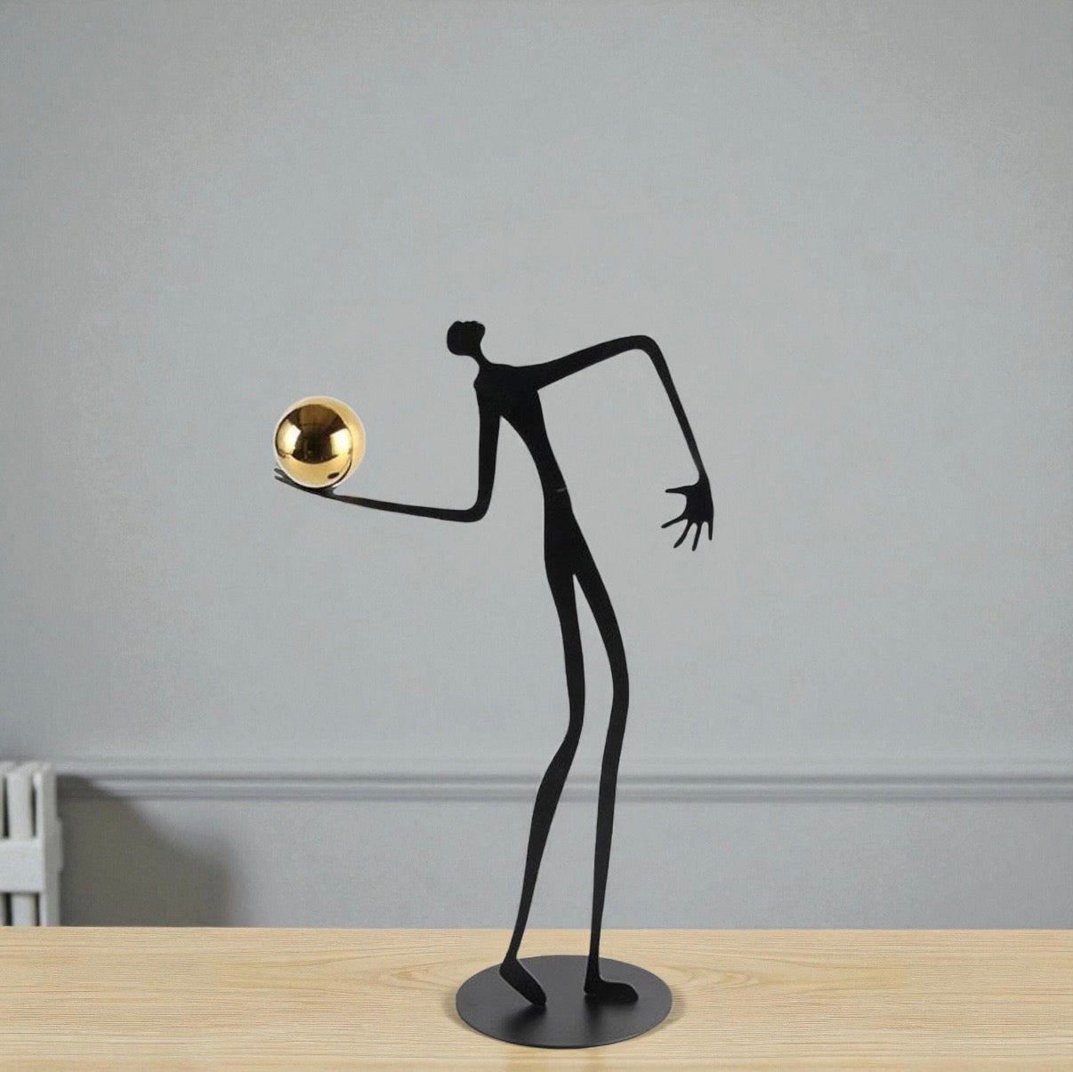 Black abstract sculpture of a figure with a gold ball on a wooden surface and gray wall background