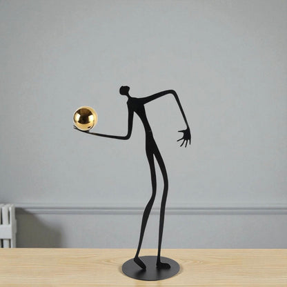 Black abstract sculpture of a figure with a gold ball on a wooden surface and gray wall background
