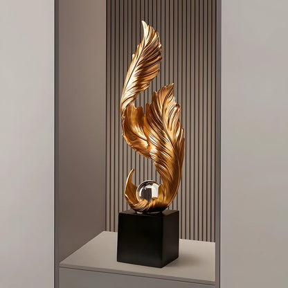 A modern decorative sculpture featuring two sweeping metallic gold leaf-like forms rising from a glossy black rectangular base, displayed on a shelf against a slatted wood and gray wall background.