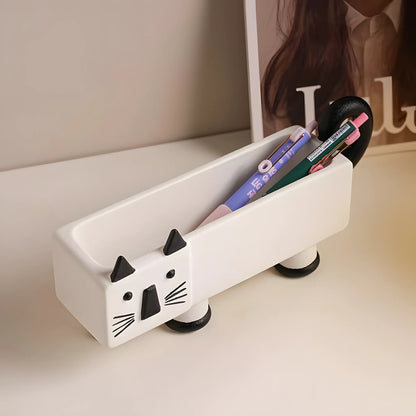 A white ceramic desk organizer shaped like a cat's body with black feet, ears, nose, and whiskers, holding pens and pencils on a light beige desk.
