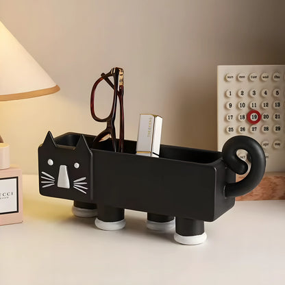 A black cat-shaped desk organizer with white facial features and white suction cup feet, holding eyeglasses and a lipstick tube, on a tabletop with a lamp and calendar in the background.