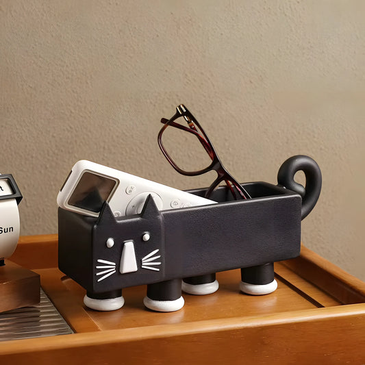 A black cat-shaped desk organizer holding eyeglasses and a remote control, placed on a wooden tray against a beige wall.