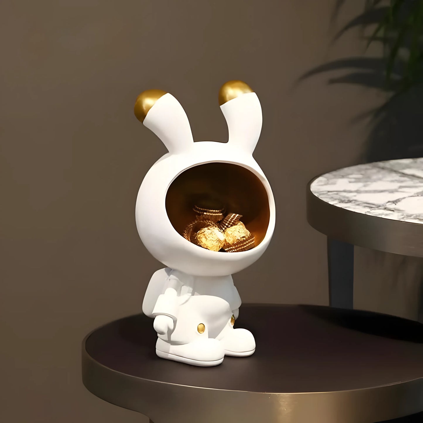 A white rabbit-shaped decorative container with gold accents sits on a dark round table, holding several wrapped chocolates inside its open head. The background is a softly lit indoor setting with a marble-topped table and a plant shadow.
