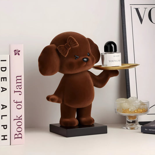 A brown plush dog figurine holding a small gold tray with a perfume bottle, displayed on a tabletop with decorative books and a framed print in the background.