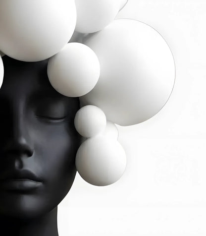 A sculptural black mannequin head with closed eyes, partially obscured by large white floating spheres against a clean white background.
