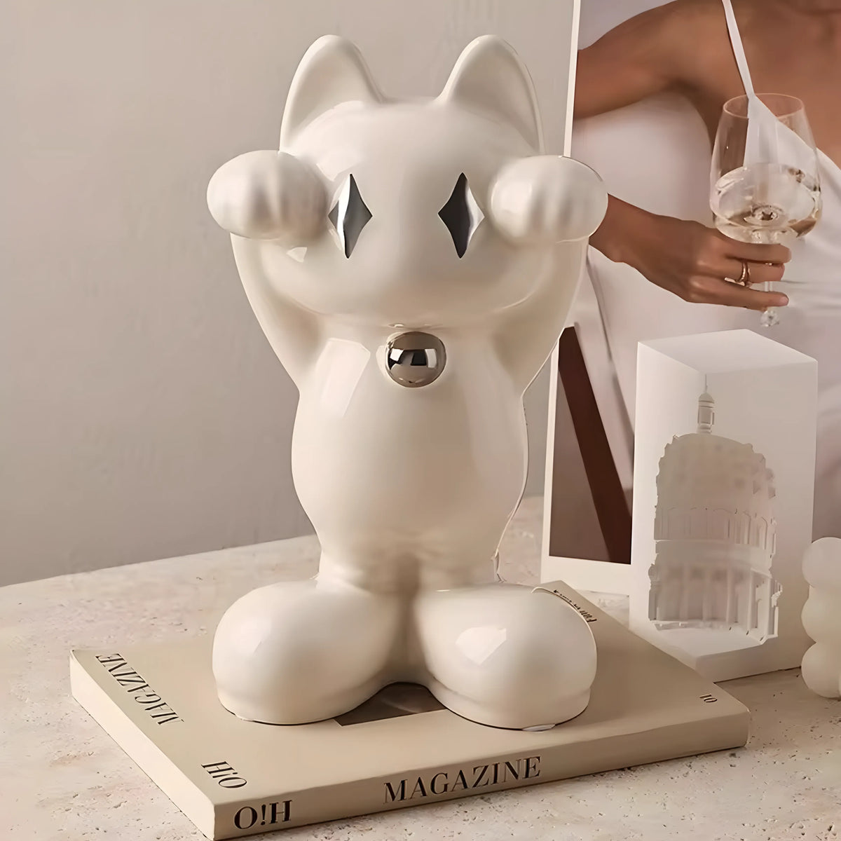 A glossy white ceramic cat-shaped decorative object with raised paws and a small metallic circular detail at the chest, displayed on a tabletop with a magazine, a glass, and a box in a styled interior setting.