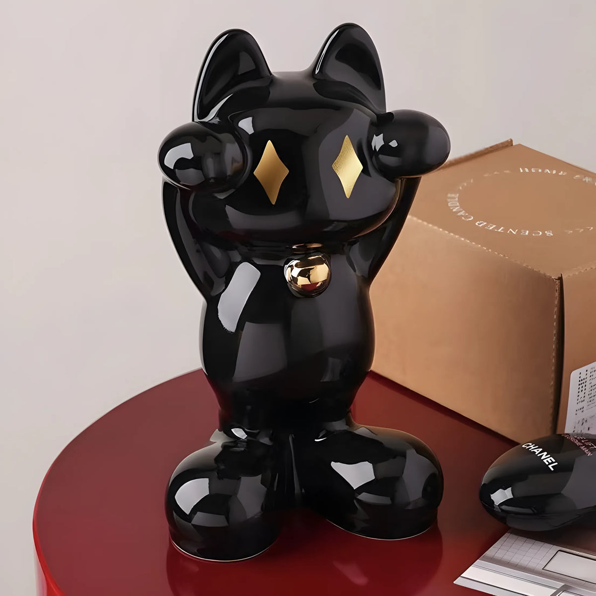 A glossy black cat-shaped decorative object with gold diamond-shaped eyes and a small gold bell at the neck, displayed on a red round table next to a brown box and a black Chanel pouch.