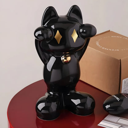 A glossy black cat-shaped decorative object with gold diamond-shaped eyes and a small gold bell at the neck, displayed on a red round table next to a brown box and a black Chanel pouch.