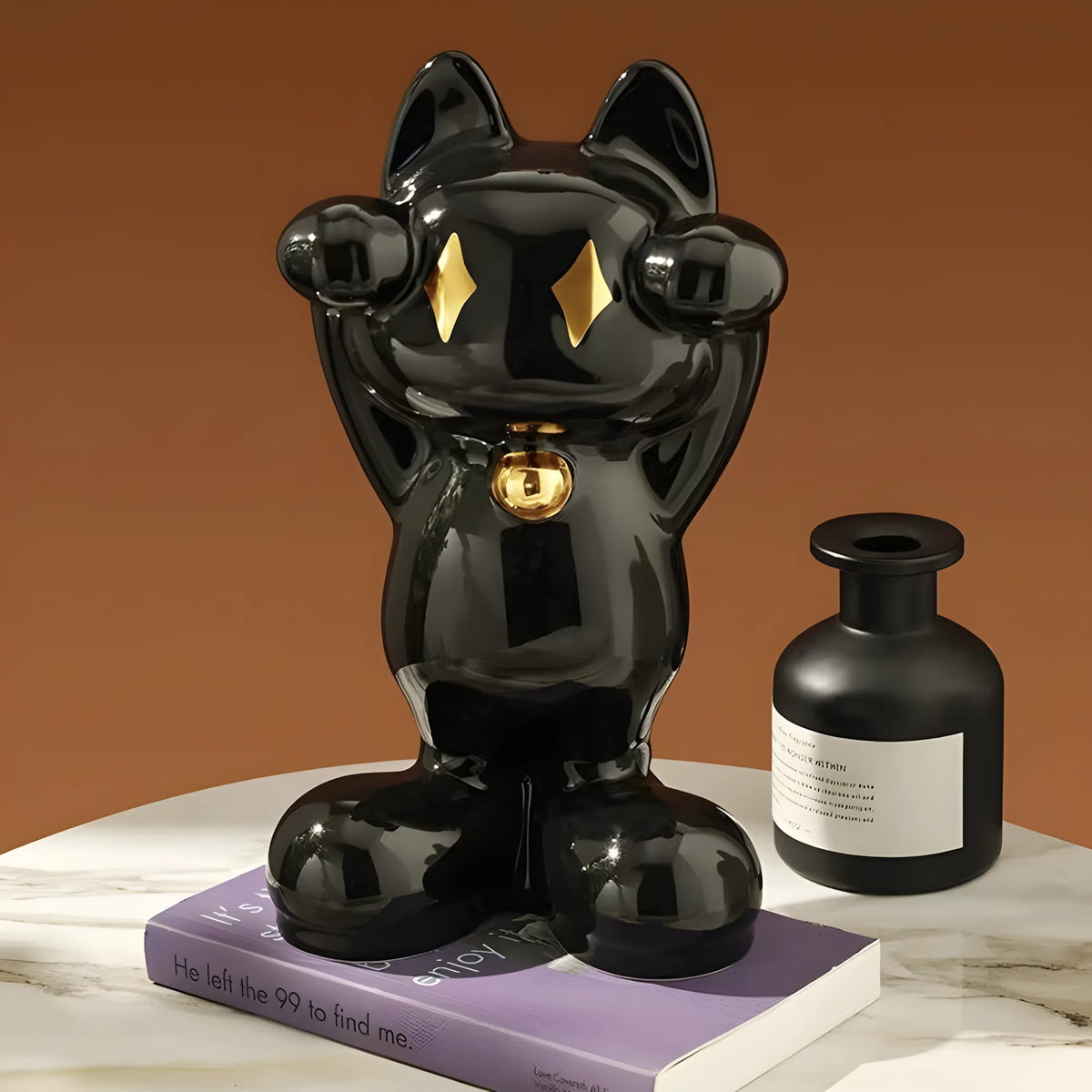 A glossy black cat figurine with gold eyes and a gold bell sits on a purple book next to a matte black bottle with a white label, set against a warm brown background.