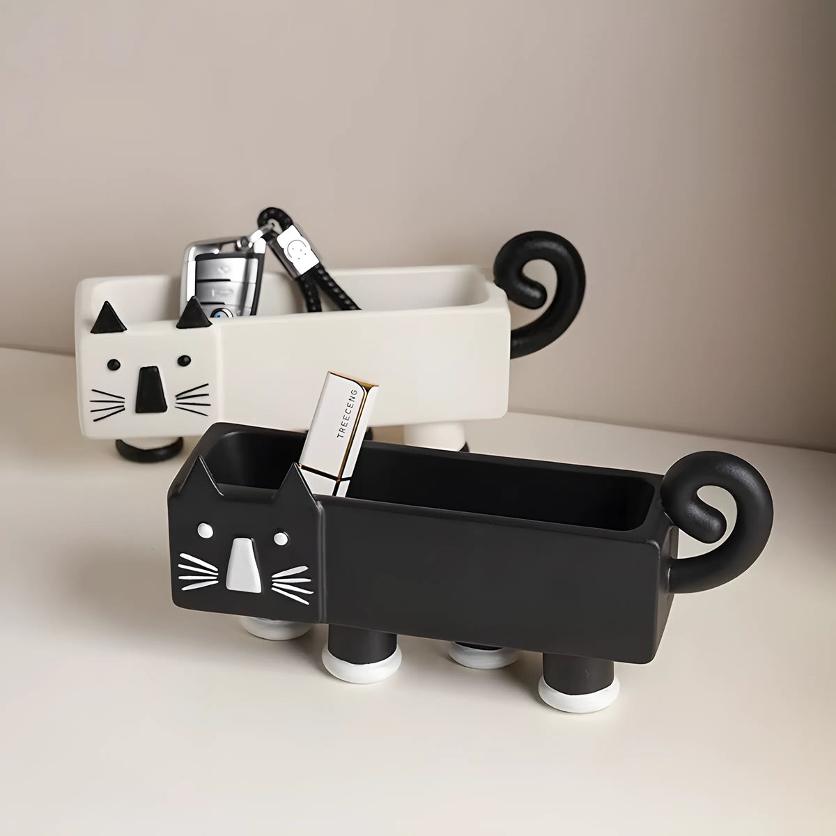 Two cat-shaped toothbrush holders on a light beige surface against a warm taupe background. The front holder is matte black with white facial details and holds a toothbrush and a cigarette pack.