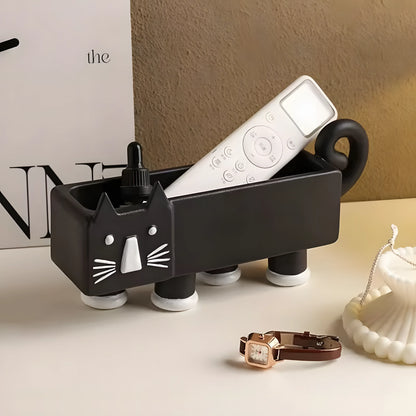 A black cat-shaped desk organizer with white accents, shown holding a white remote control and a small black bottle. The organizer has a cat face on the front, four legs, and a handle on the back.