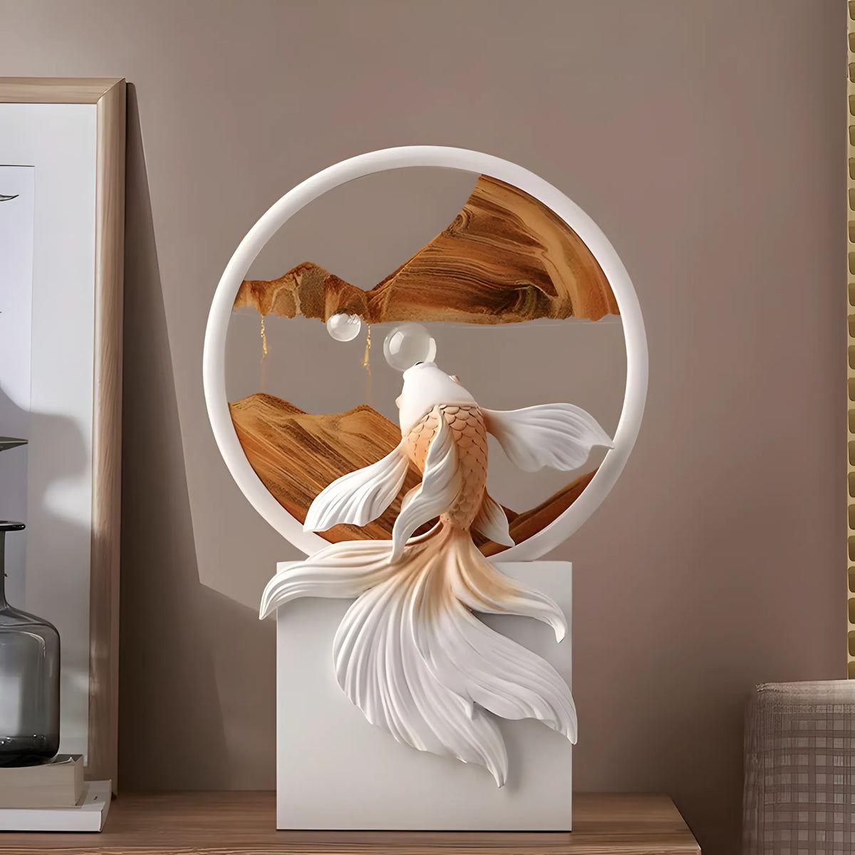 A decorative white fish sculpture with flowing fins, set inside a circular frame with layered wood panels, displayed on a white pedestal against a warm taupe wall.