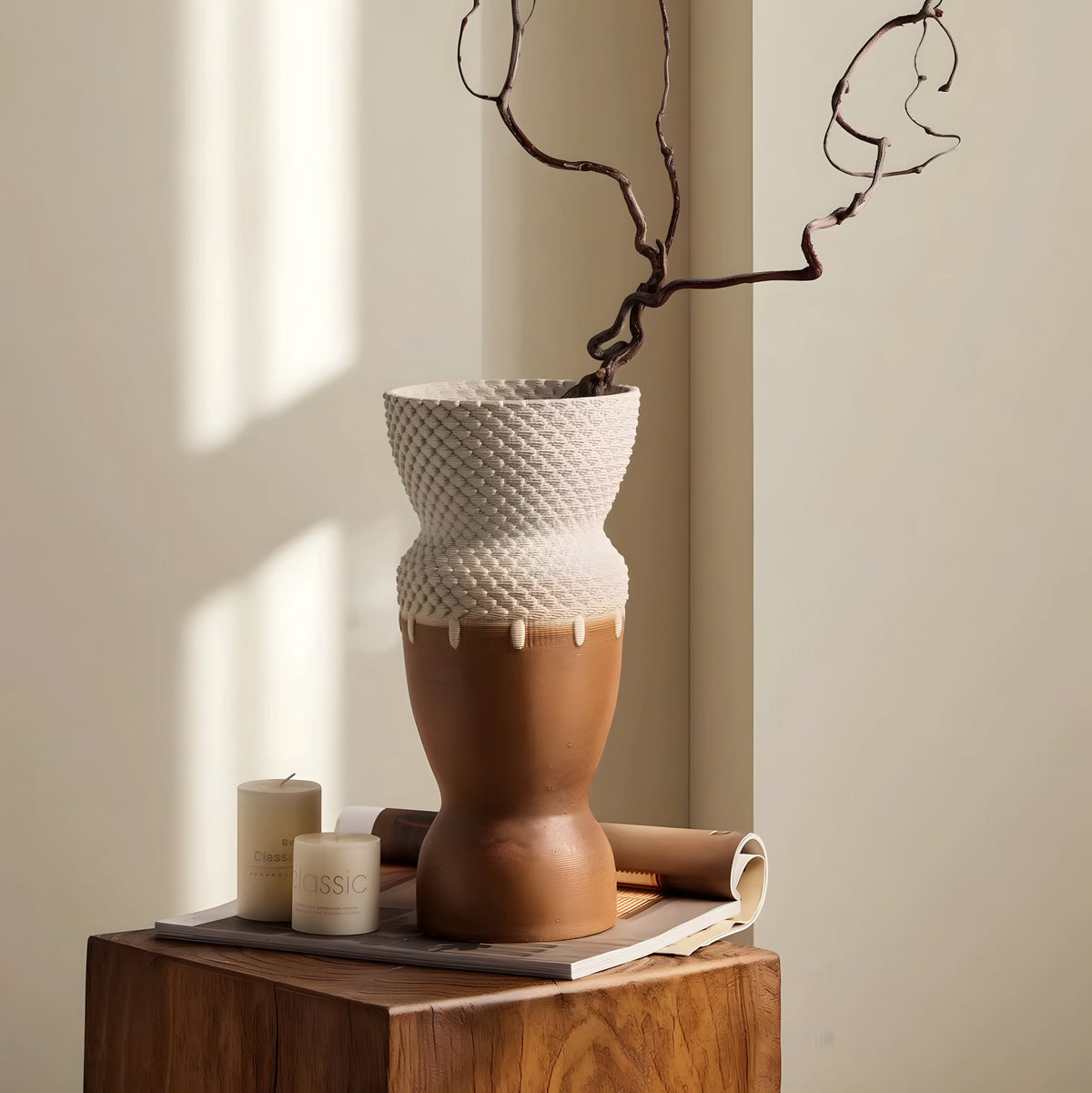 A sculptural ceramic vase with a textured cream upper section and a smooth terracotta lower section, displayed on a wooden block with candles and a rolled paper in a warm neutral room.