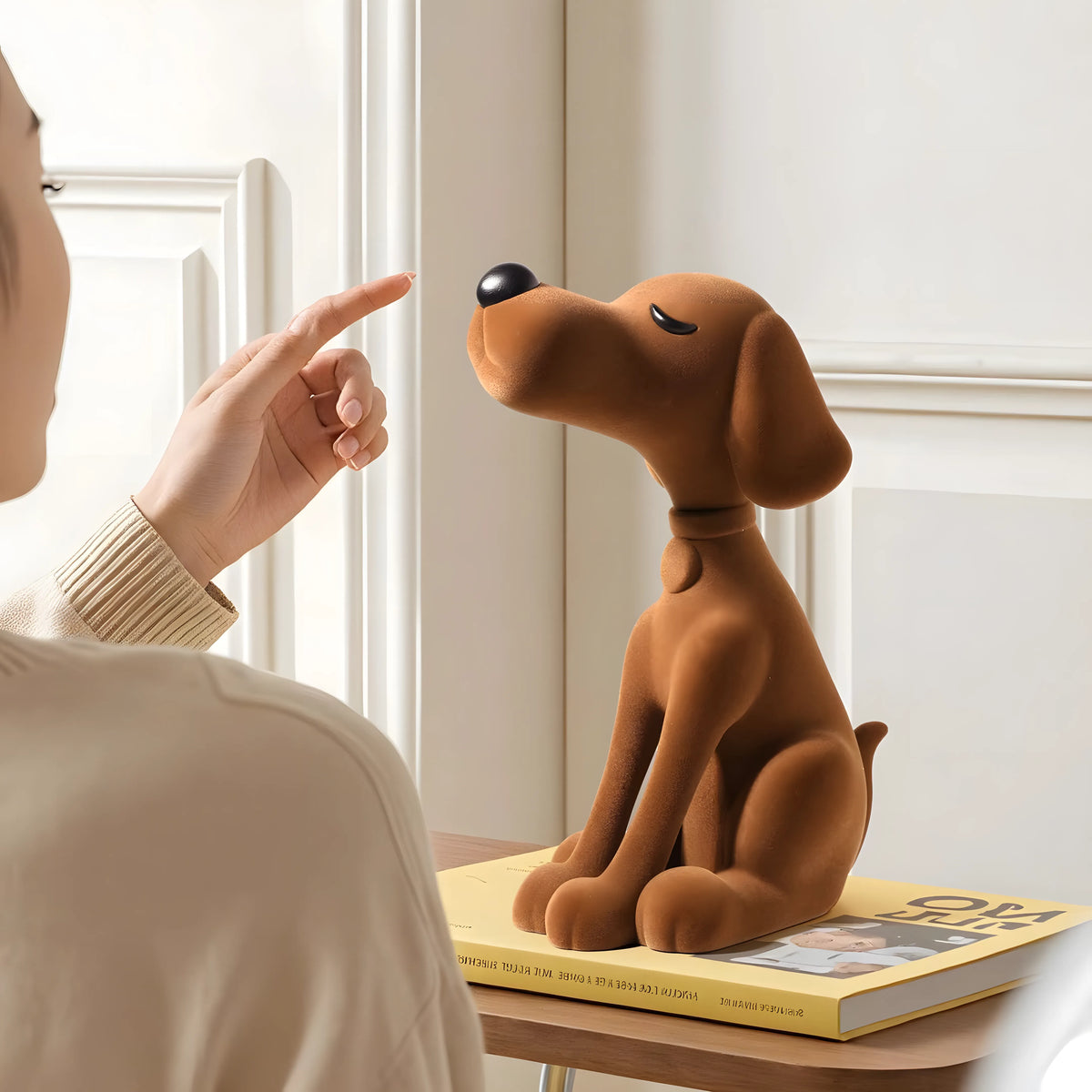 A brown dog-shaped figurine sits on a yellow book on a table, with a person pointing at it in a bright indoor setting.