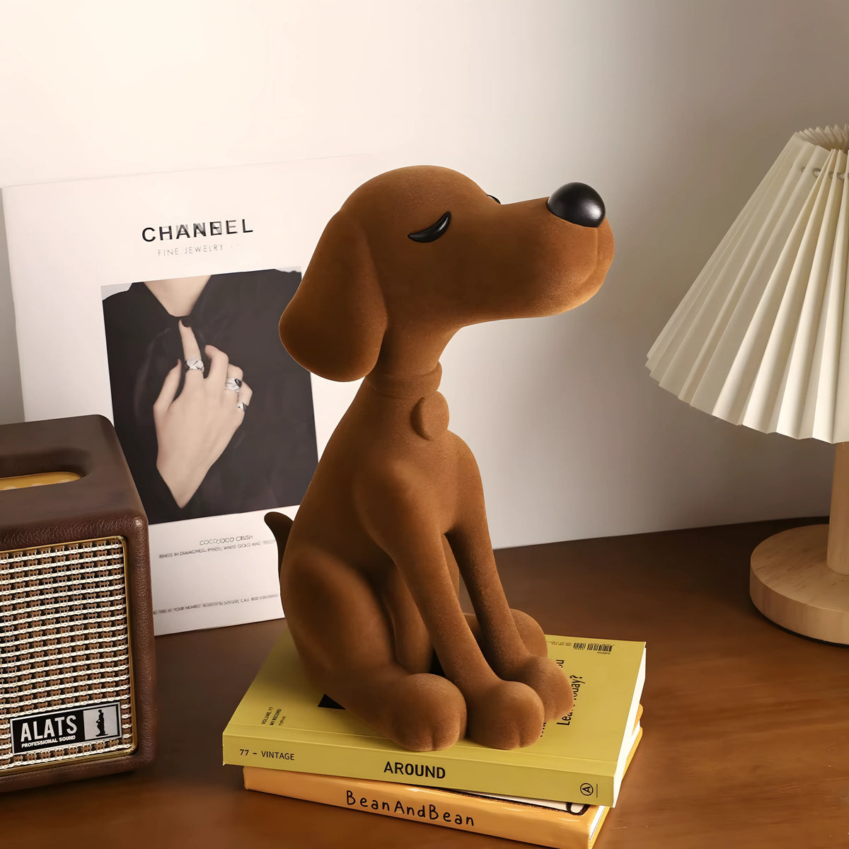 A brown plush dog figurine sitting on a stack of yellow books on a wooden table, styled with a vintage radio, a Chanel magazine, and a table lamp in the background.