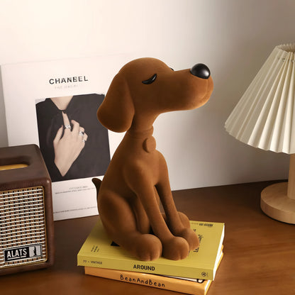 A brown plush dog figurine sitting on a stack of yellow books on a wooden table, styled with a vintage radio, a Chanel magazine, and a table lamp in the background.