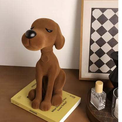 A brown plush dog figurine sitting on a yellow book on a wooden table, with a Loewe perfume bottle and a framed black-and-white geometric print in the background.
