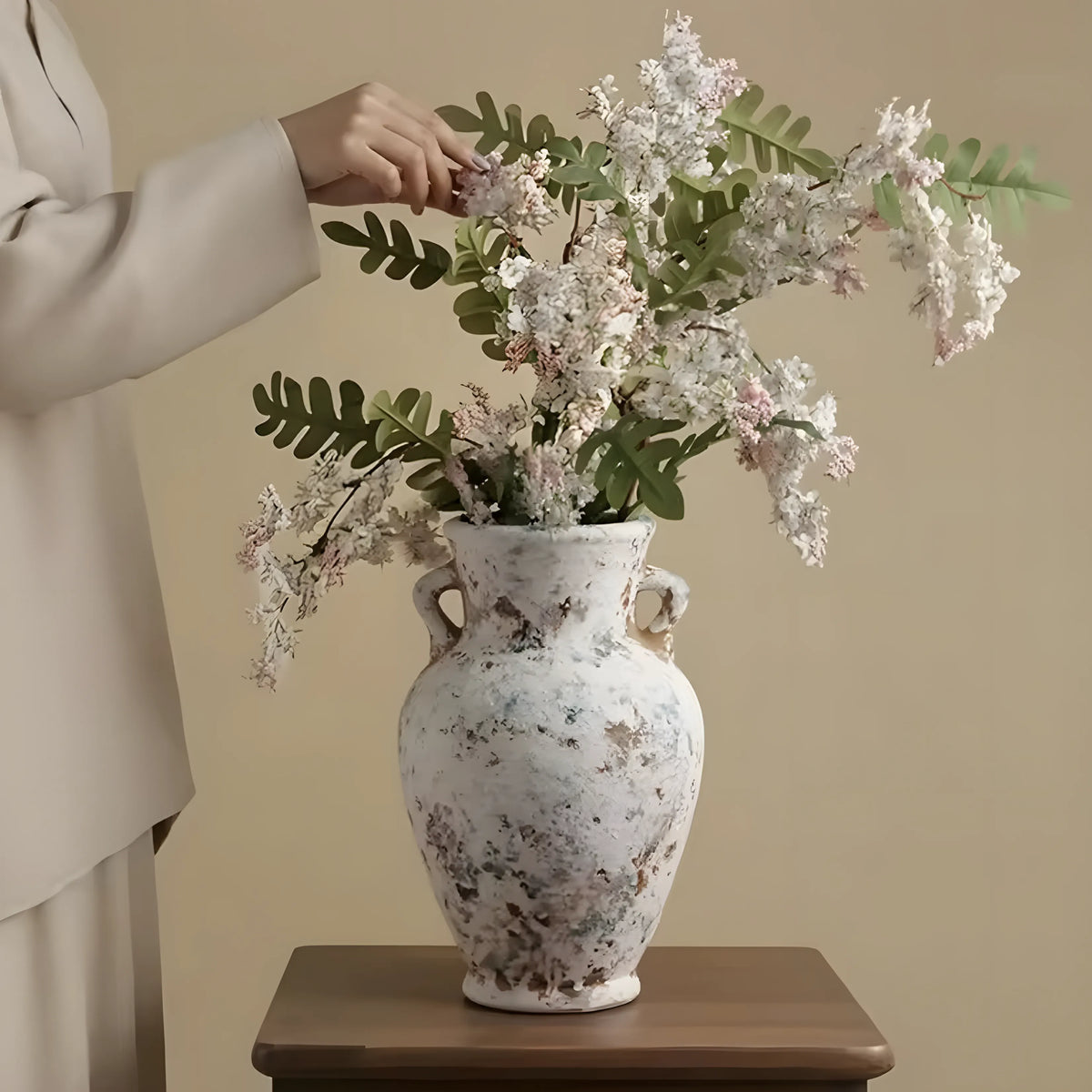 A person in a light beige outfit adjusts a large white ceramic vase filled with white blossoms and green leaves, placed on a wooden table against a warm beige wall.