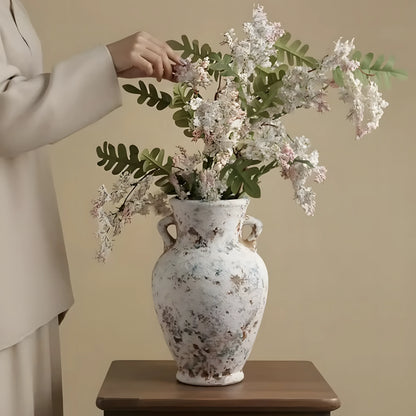 A person in a light beige outfit adjusts a large white ceramic vase filled with white blossoms and green leaves, placed on a wooden table against a warm beige wall.