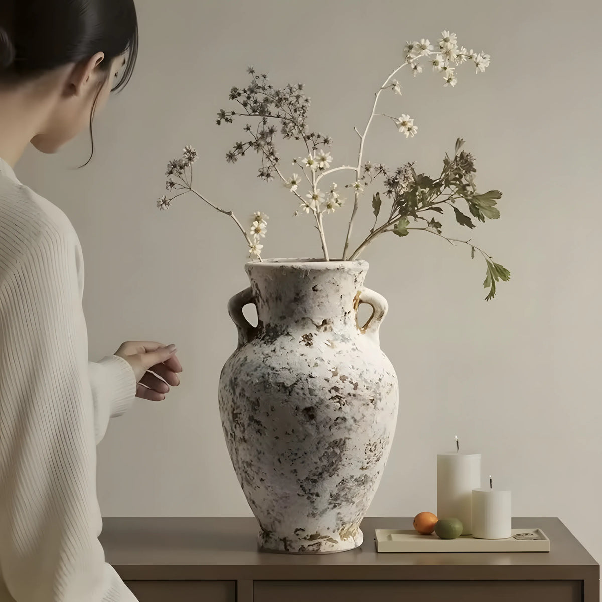 A large distressed ceramic vase with two handles sits on a wooden table, filled with dried white flowers and green stems. A woman in a white ribbed sweater is partially visible on the left, and two white candles with small fruit are on the right against a plain beige wall.