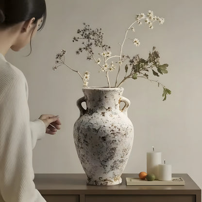 A large distressed ceramic vase with two handles sits on a wooden table, filled with dried white flowers and green stems. A woman in a white ribbed sweater is partially visible on the left, and two white candles with small fruit are on the right against a plain beige wall.