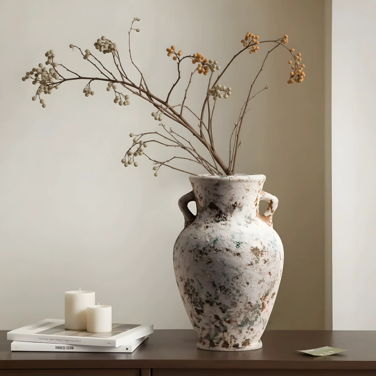 A large distressed ceramic vase with two side handles, filled with dried branches and small yellow flowers, displayed on a dark wooden table next to two white candles and a stack of books.