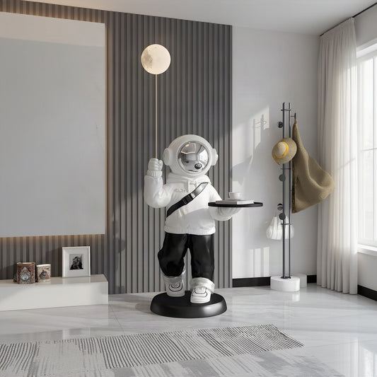 A whimsical astronaut-shaped floor lamp or decorative statue stands on a round black base in a modern interior. The figure is mostly white with black pants and a black shoulder strap, holding a thin pole with a spherical moon-shaped bulb in one hand and a small tray in the other.