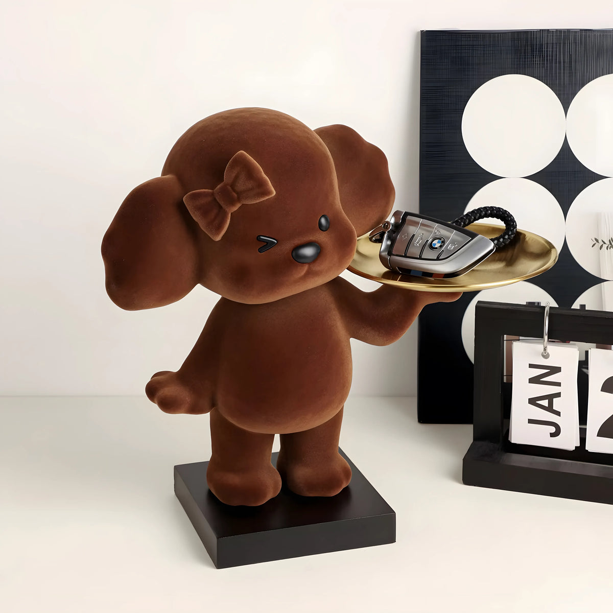 A brown plush puppy figurine holding a gold tray with a car key, displayed on a black base next to a black and white calendar and a black-and-white circular wall art piece.