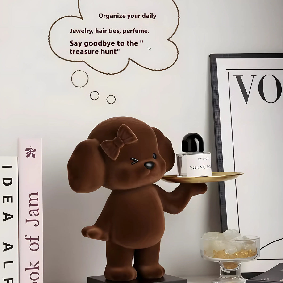 A brown plush dog figurine holding a small tray with a perfume bottle, displayed on a tabletop with decorative books, a framed print, and a thought bubble graphic on a white wall.