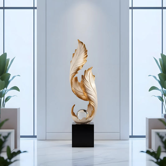 A modern abstract gold sculpture resembling stylized leaves or flowing forms, mounted on a black rectangular pedestal in a bright, minimalist interior with large windows and potted plants.