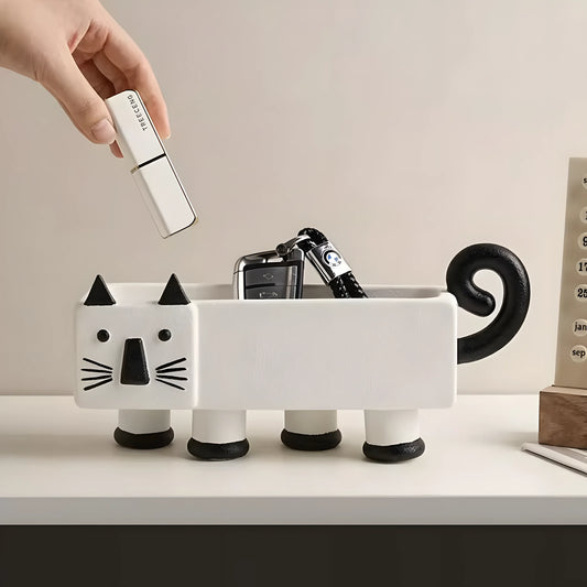 A white ceramic cat-shaped desk organizer with black accents, shown on a white desk with a hand placing a white USB drive into its open belly. The cat has black feet, a curled black tail, and black facial features.