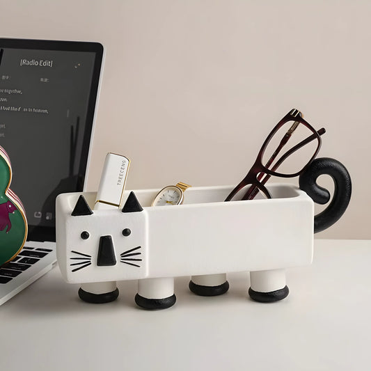 A white ceramic cat-shaped desk organizer with black accents, holding eyeglasses, a watch, and a small pen, placed on a desk next to a laptop.