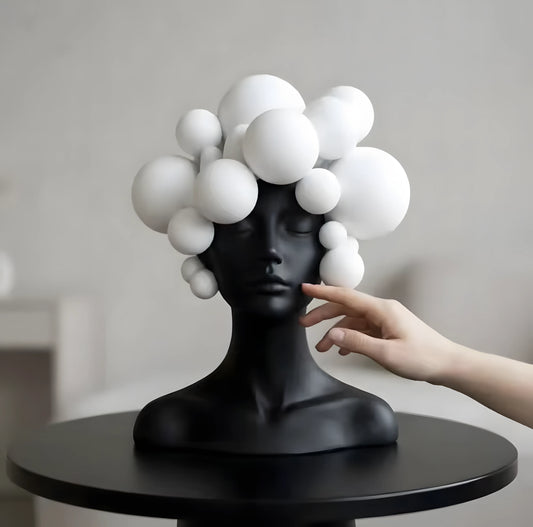 A matte black female bust sculpture with a cluster of white spherical shapes forming her hair, displayed on a round black table with a human hand reaching toward it.