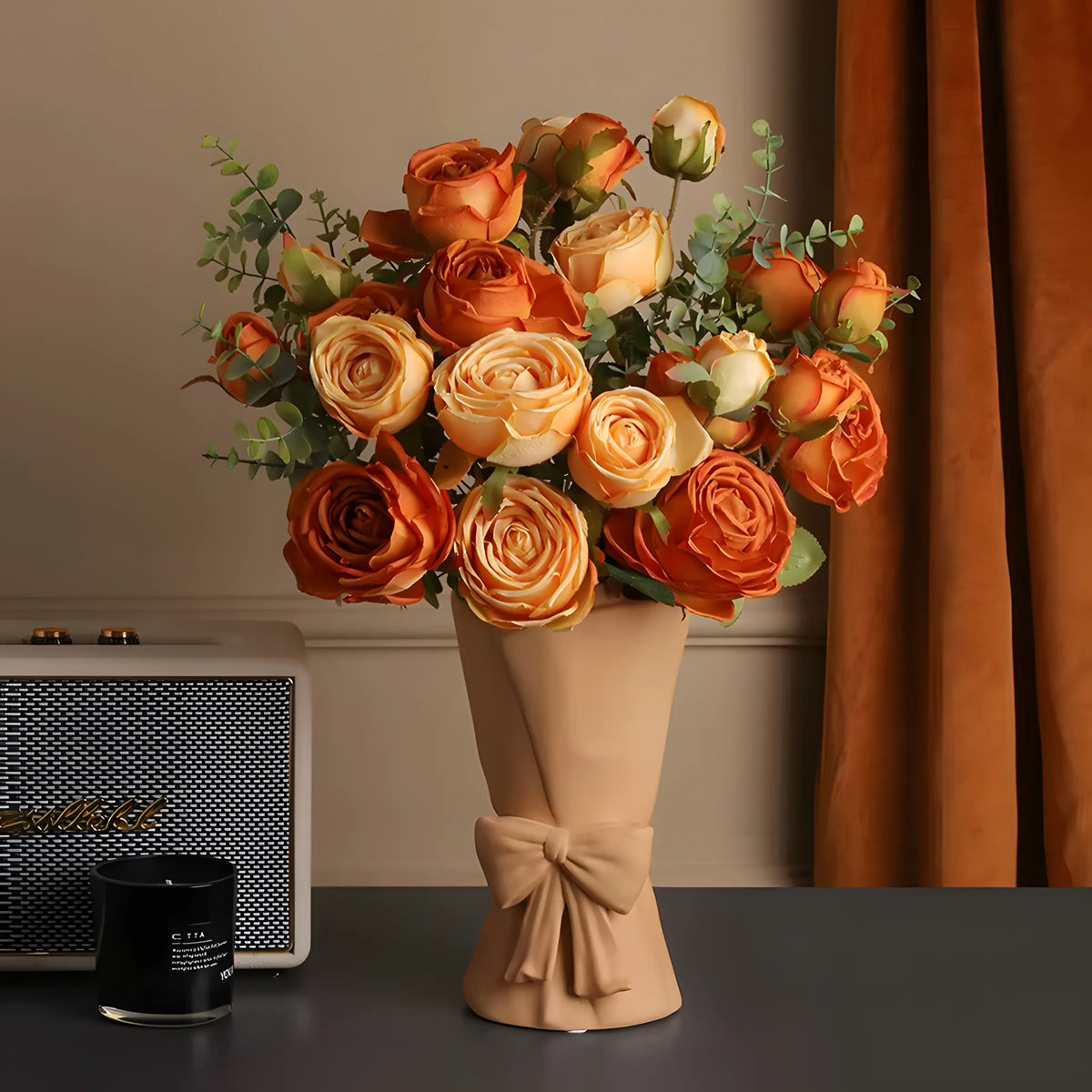 A bouquet of artificial orange and peach roses with green foliage, wrapped in a beige paper sleeve tied with a bow, displayed on a dark tabletop with a curtain and a speaker in the background.
