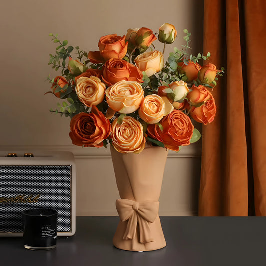 A bouquet of artificial orange and peach roses with green foliage, wrapped in a beige paper sleeve tied with a bow, displayed on a dark tabletop with a curtain and a speaker in the background.