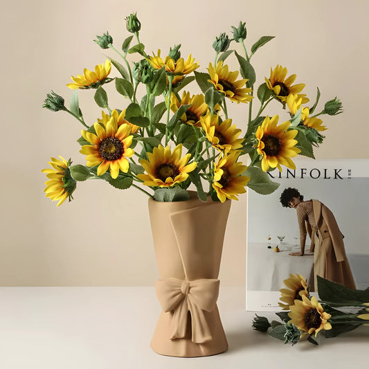 A bouquet of artificial yellow sunflowers with green stems and buds, arranged in a beige fabric vase with a bow, set on a light surface against a warm beige background. A magazine and a few loose sunflowers are placed beside it.