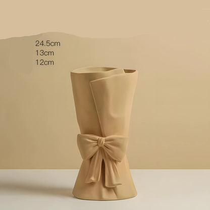 A beige ceramic flower holder shaped like a rolled sheet of paper with a bow at the base, shown on a matching beige background with black text listing dimensions.