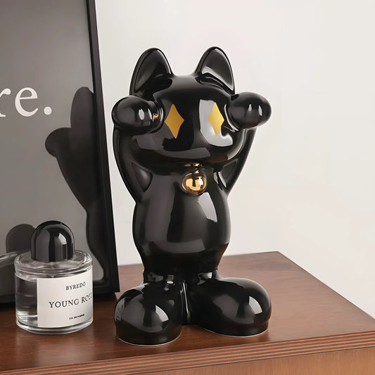 A glossy black cat figurine with gold diamond-shaped eyes and a gold bell around its neck, standing on a wooden table next to a perfume bottle and a framed print.