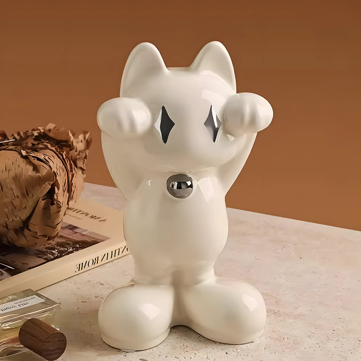 A glossy white ceramic cat figurine with raised paws and diamond-shaped cutout eyes, displayed on a tabletop with a warm brown background and styled props.