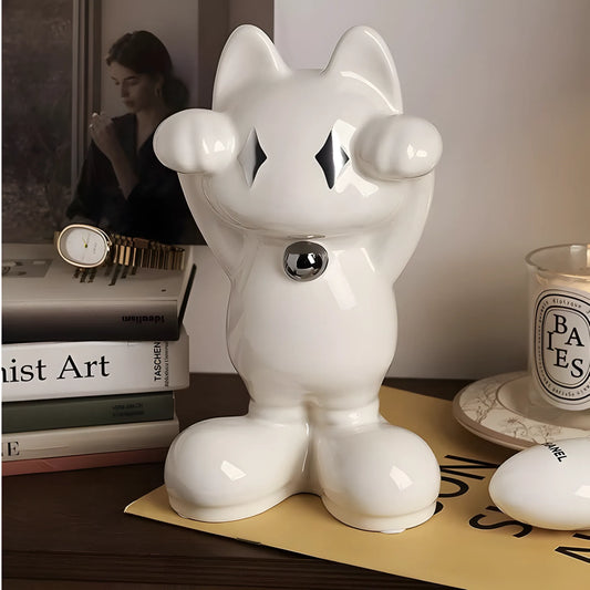 A glossy white ceramic cat figurine with raised paws and a small metallic silver bell at the neck, displayed on a tabletop with books, a candle, and a watch in a styled interior setting.