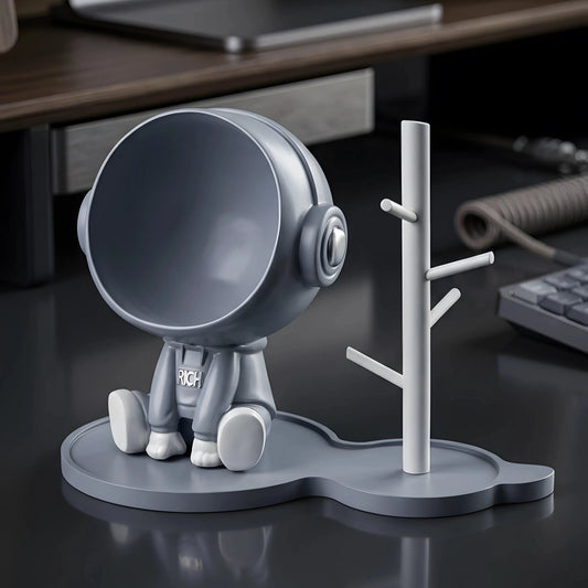 A modern desk organizer set featuring a small astronaut-shaped phone stand and a matching tree stand, both in matte gray and white, displayed on a dark reflective desk surface with a blurred office background.