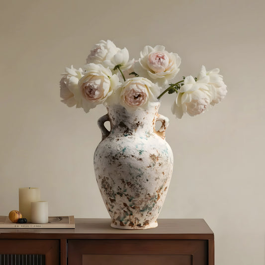 A large distressed ceramic vase with two side handles, filled with large white and blush pink peony flowers, sits on a dark wood sideboard against a warm beige wall. A small candle and book are visible on the left.